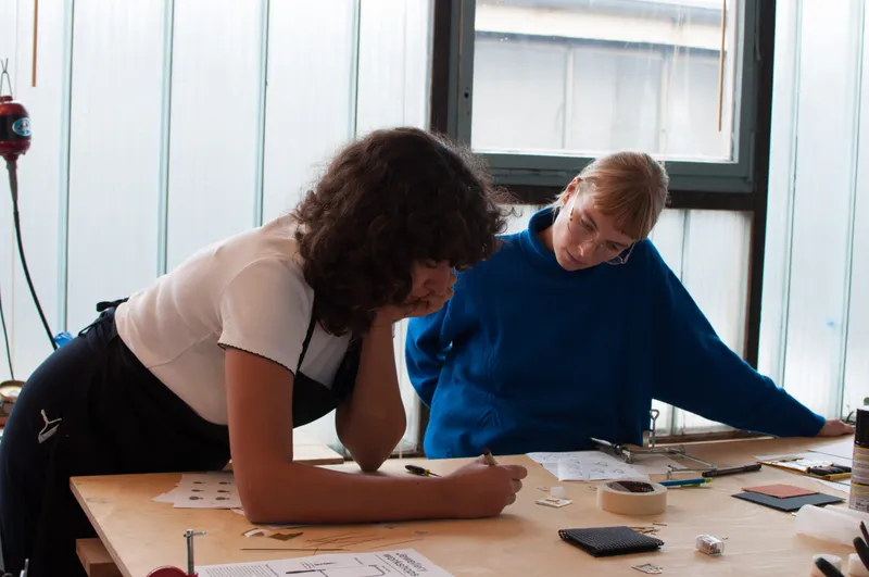 Two participants sketching pendant designs on paper at a plywood workbench spread with files, saws, tape and metal offcuts