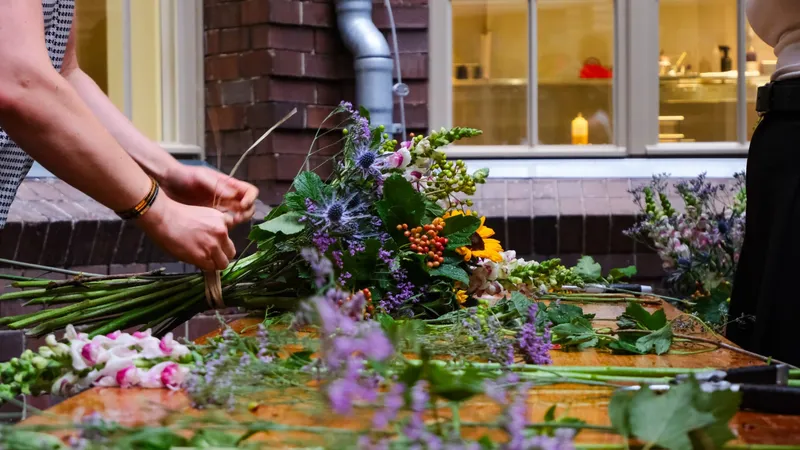 Hands tying a seasonal bouquet of sunflowers, snapdragons and thistle at an outdoor wooden table covered in cut stems