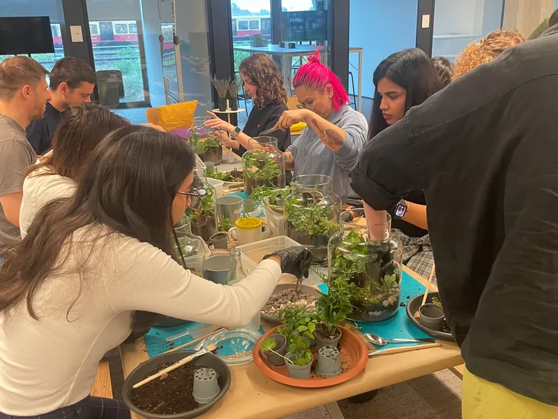 A group leaning over a crowded worktable of glass jars, seedlings and moss while a facilitator points out a planting step