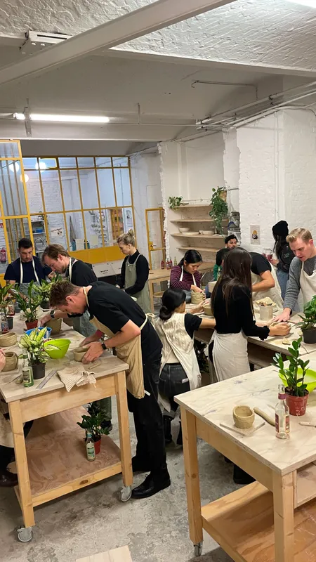 A full studio of aproned participants shaping clay pots at plywood worktables lined with houseplants and tools