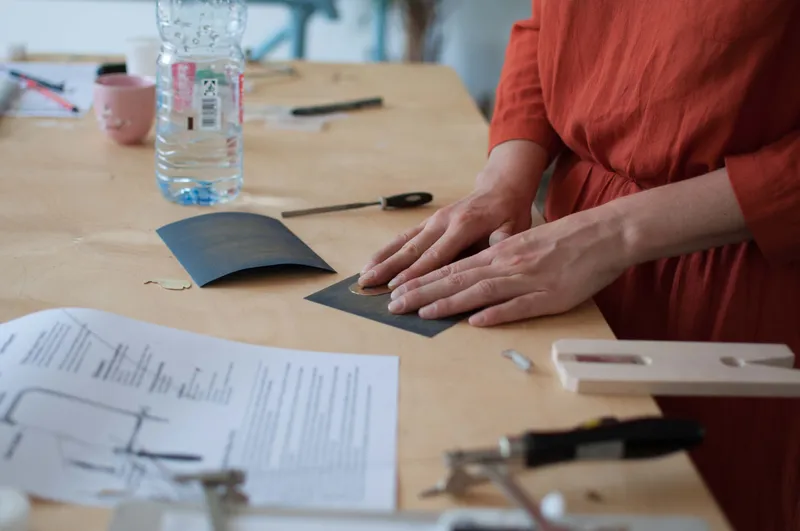 Hands sanding a small brass pendant on emery paper at a workbench with hand files, instruction sheets and a bench pin