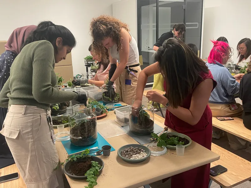 Colleagues in gloves planting glass jar terrariums at long office tables laid out with soil trays, gravel bowls and tools