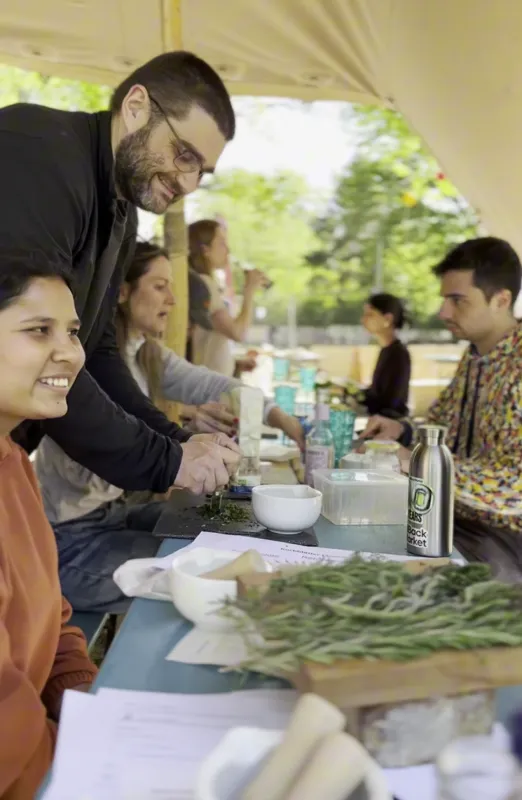 Participants grinding herbs with pestles at a long outdoor table under a canopy while a facilitator leans in to help