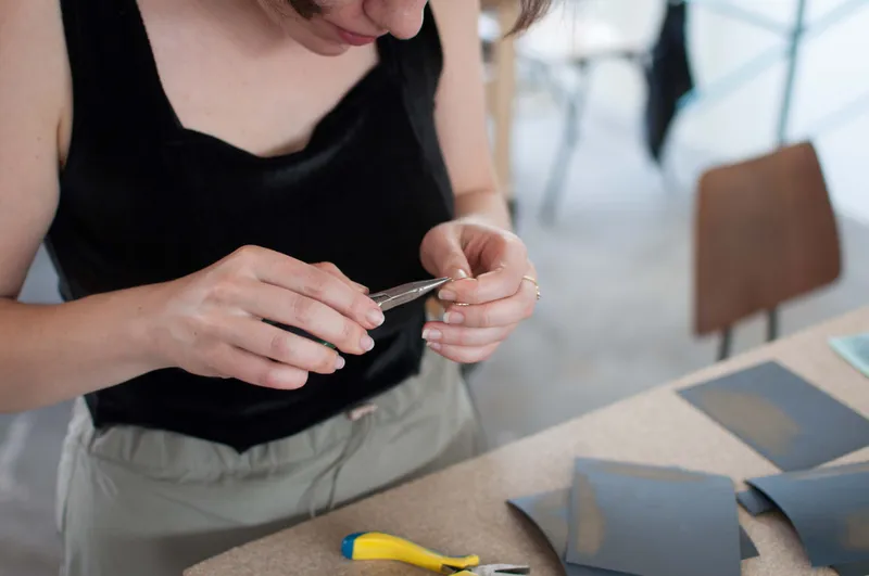 A participant shaping a metal pendant with round-nose pliers beside cut sheets of emery paper and a pair of flat pliers