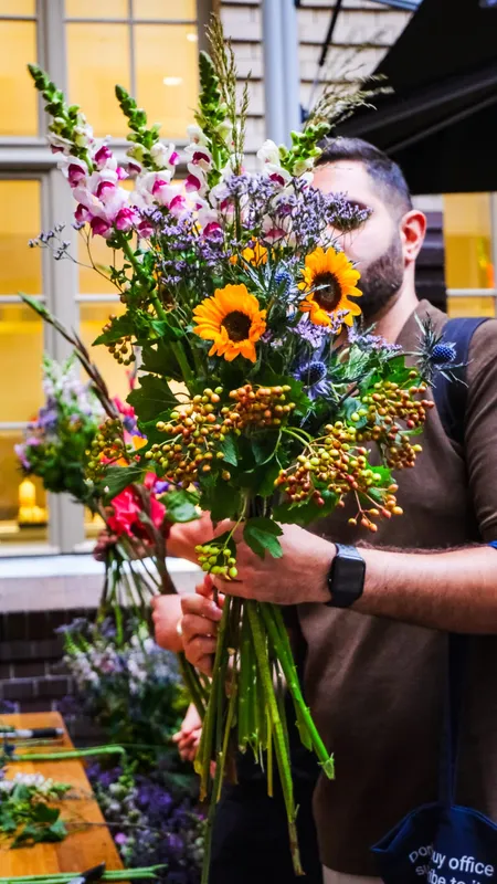 A participant holding up a finished bouquet of sunflowers, snapdragons and berry branches at a courtyard flower bar
