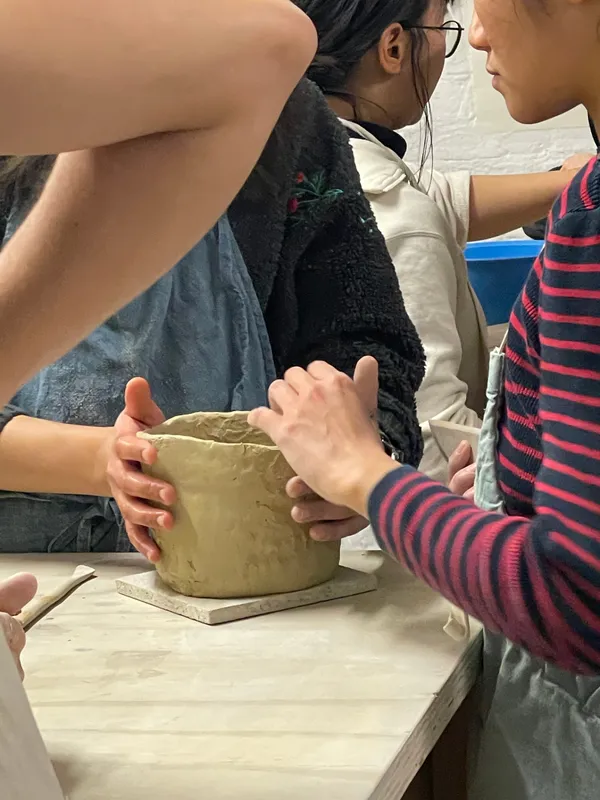 Close-up of hands pressing and shaping a wet clay planter on a board while others work alongside