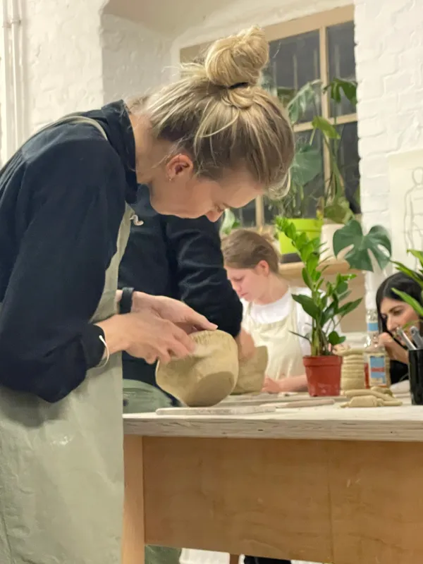 A participant smoothing the walls of a hand-built clay pot at a worktable surrounded by plants and drying pieces