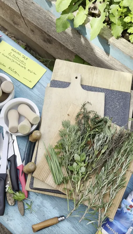 Freshly cut rosemary, sage, thyme and chives laid out on wooden boards beside mortars, knives and a herb salt recipe card on a blue garden table